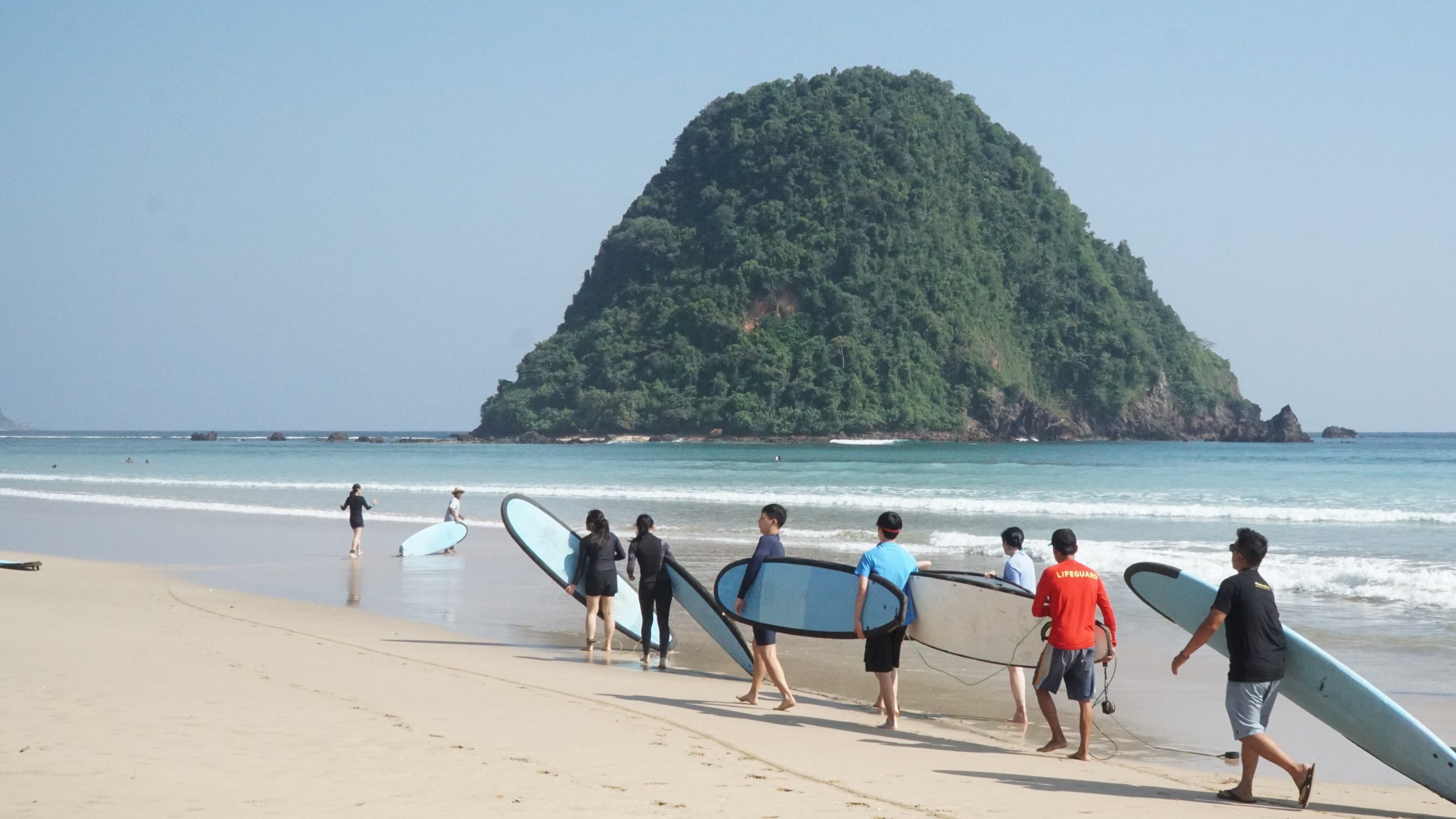 Students carrying surfboards along the beach beside Banyuwangi's iconic rock island